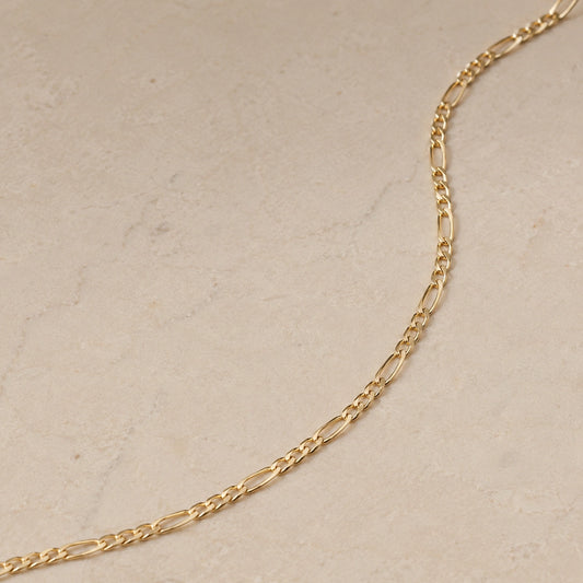 Close up of a 14ct gold vermeil Figaro chain necklace laid on a light beige stone background, showing the elongated and round link pattern.