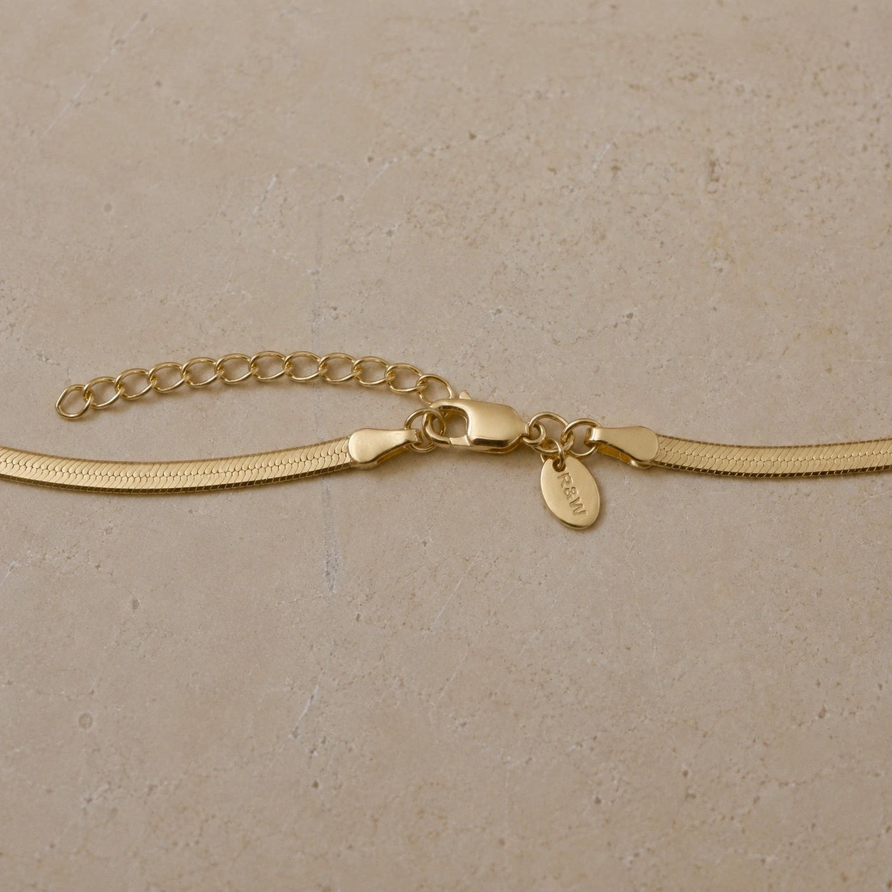 Close-up of a 14ct gold vermeil snake chain necklace featuring a lobster clasp, extender chain and Rose & Wolf branded tag on a neutral stone background.