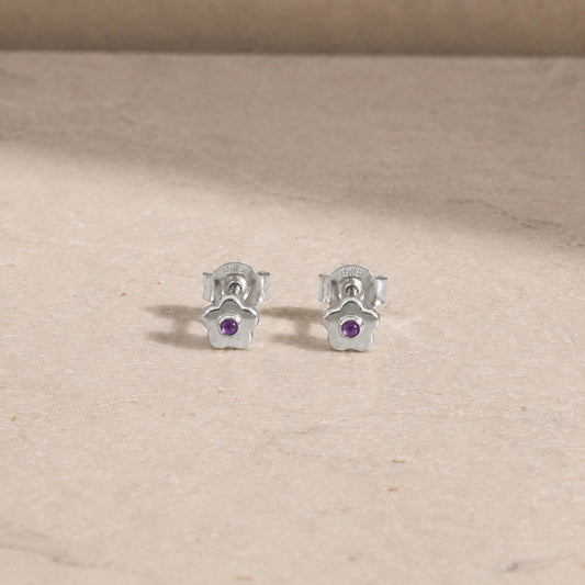 Pair of sterling silver flower stud earrings with amethyst centre stones, displayed on a light beige stone surface with 925 butterfly backs visible.