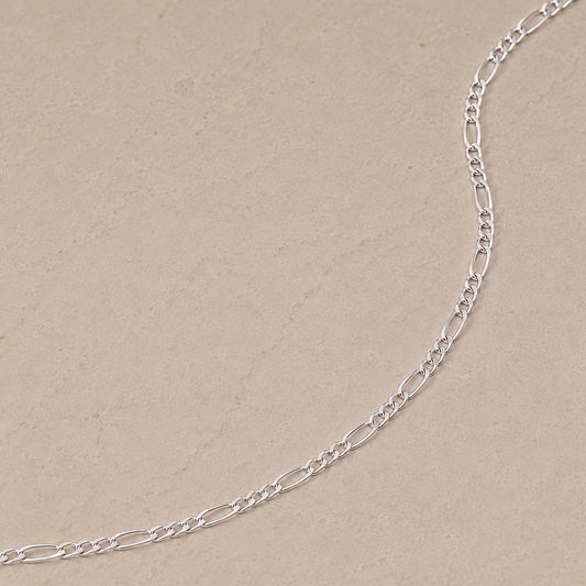 Close-up of a sterling silver Figaro chain necklace with alternating long and short links on a neutral beige stone background.