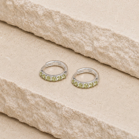 Sterling silver peridot huggie hoop earrings with five light green gemstones on each hoop, displayed on a textured cream stone surface with visible 925 hallmark inside each earring.