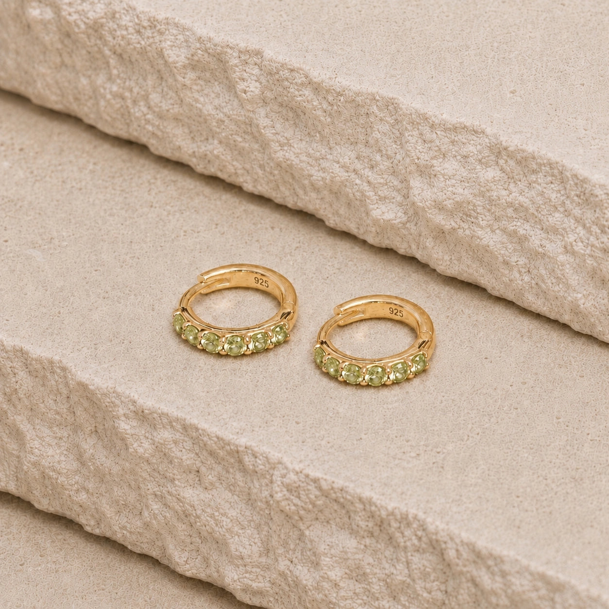 Pair of 14ct gold vermeil huggie hoop earrings with faceted peridot gemstones on a textured beige stone surface, featuring a visible 925 hallmark on the inner band.