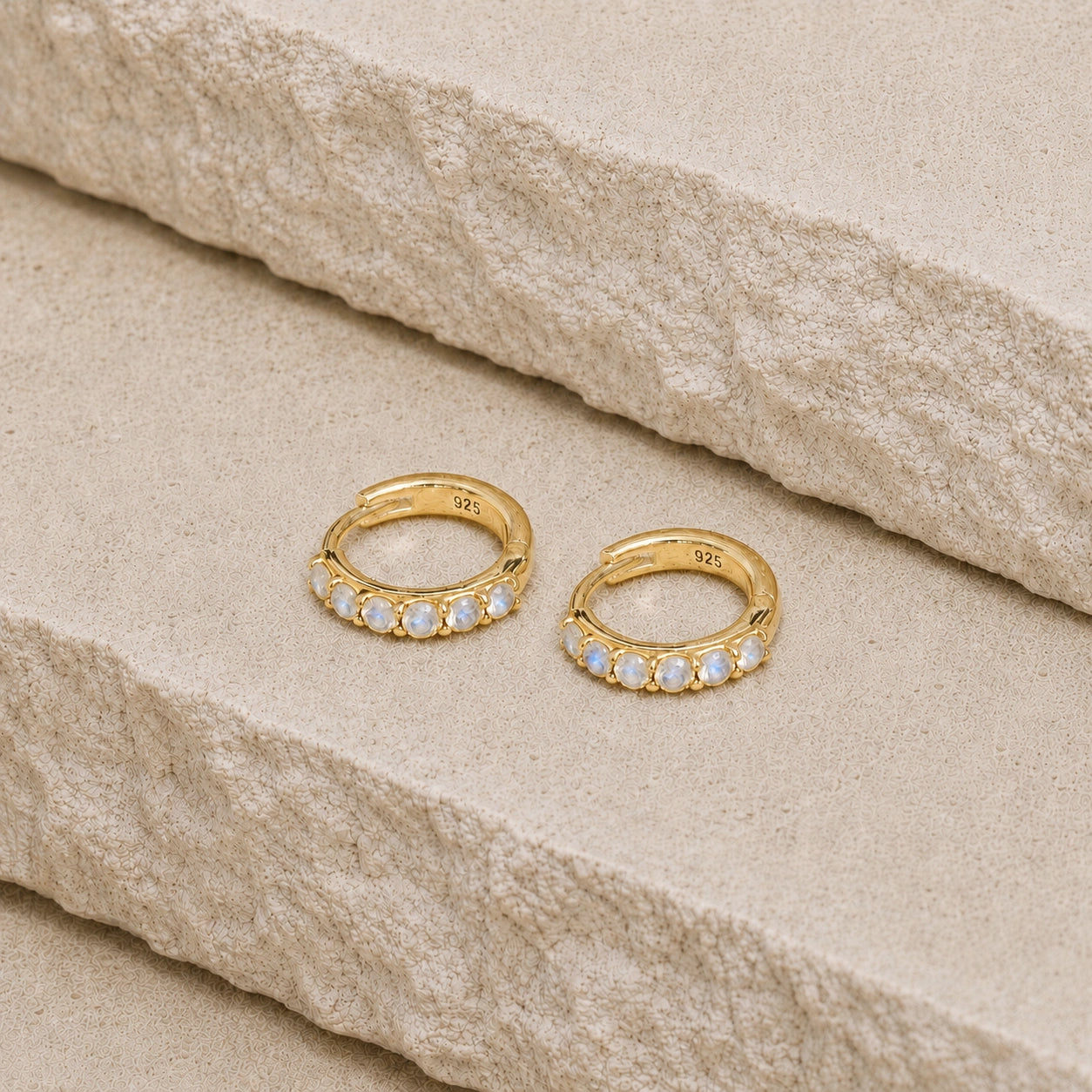 Pair of 14ct gold vermeil rainbow moonstone huggie hoop earrings displayed on a textured beige stone surface, featuring six round rainbow moonstones on each hoop and a 925 stamp on the inner hoops.