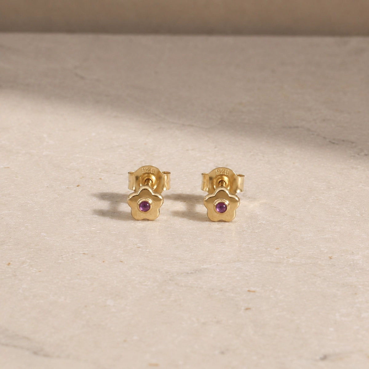Pair of 14ct gold vermeil flower stud earrings with amethyst centre stones, displayed on a light beige stone surface with 925 butterfly backs visible.