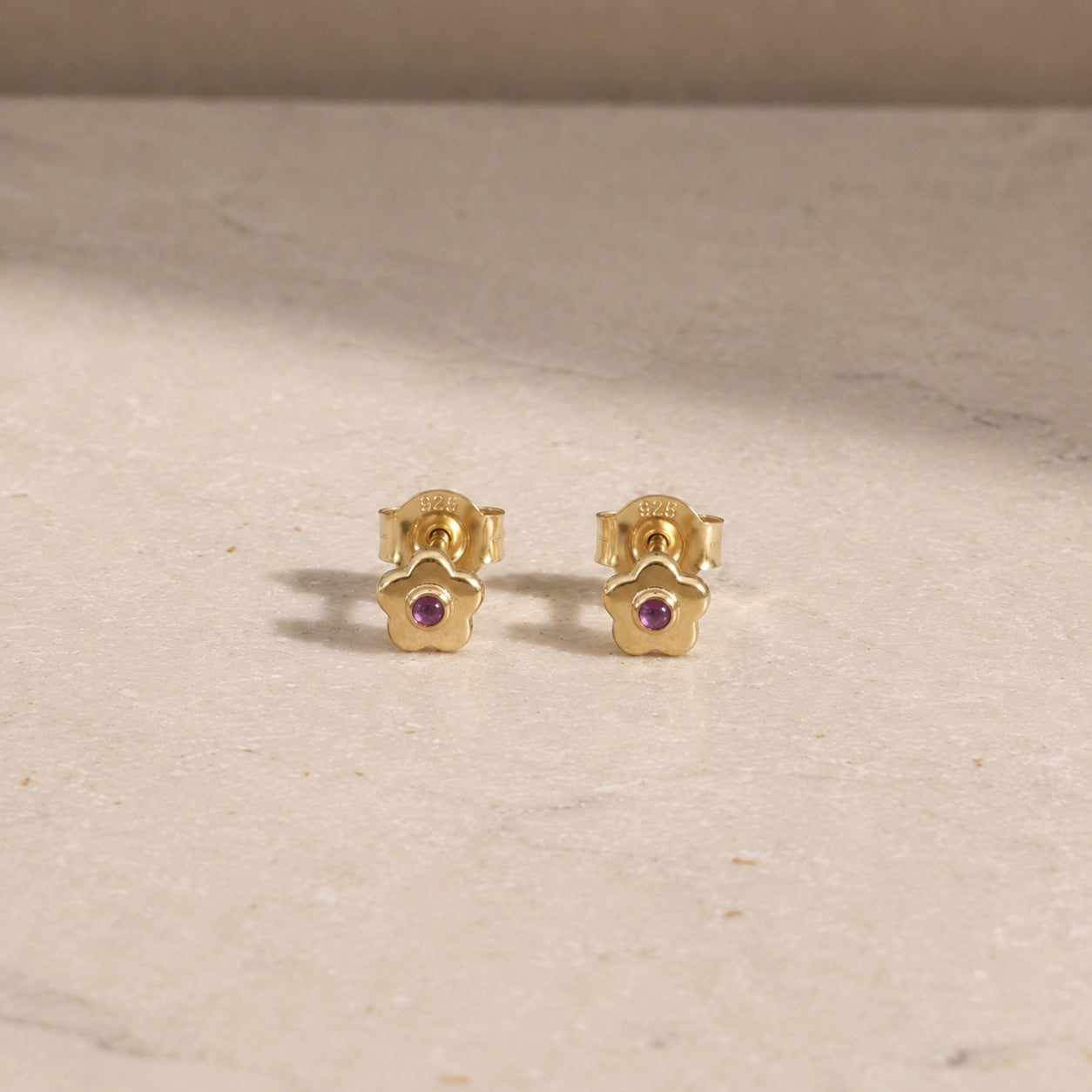 Pair of 14ct gold vermeil flower stud earrings with amethyst centre stones, displayed on a light beige stone surface with 925 butterfly backs visible.