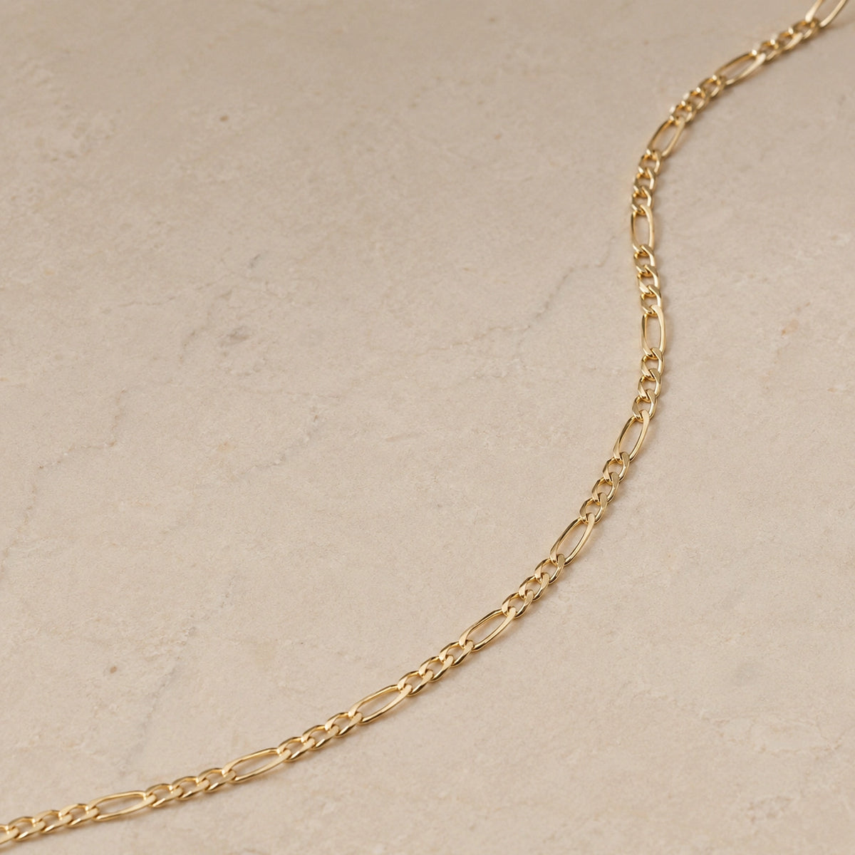 Close up of a 14ct gold vermeil Figaro chain necklace laid on a light beige stone background, showing the elongated and round link pattern.