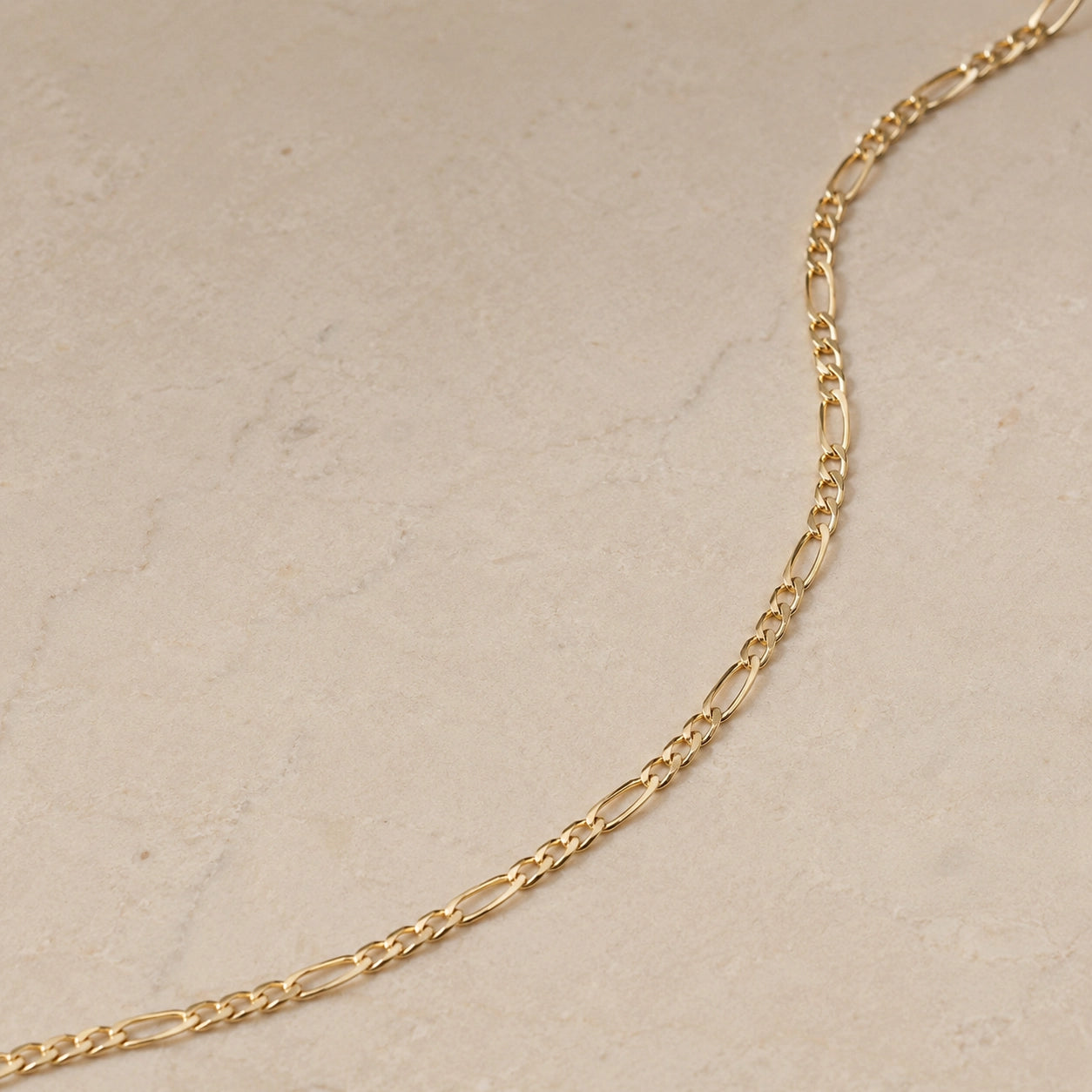 Close up of a 14ct gold vermeil Figaro chain necklace laid on a light beige stone background, showing the elongated and round link pattern.
