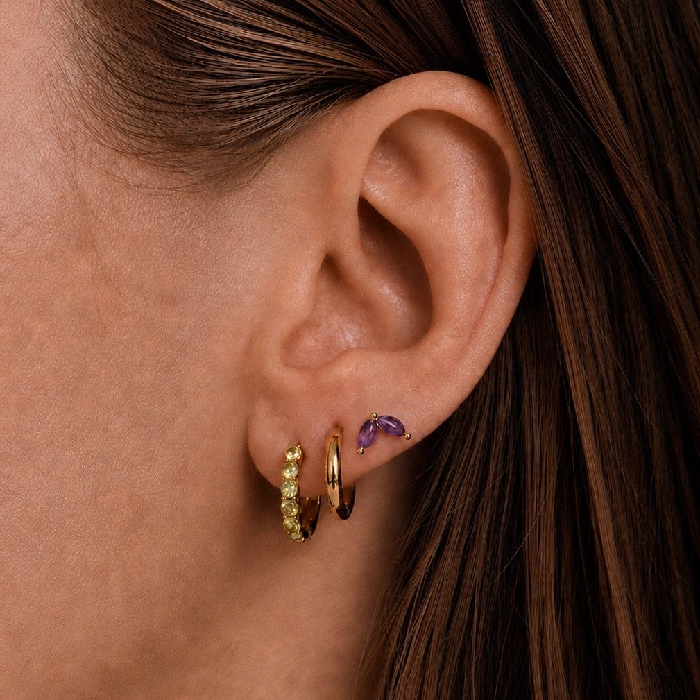 Close up of a model wearing 14ct gold vermeil faceted peridot huggie earrings and amethyst marquise stud earrings, shown on the ear with natural skin texture and brown hair tucked behind the ear.