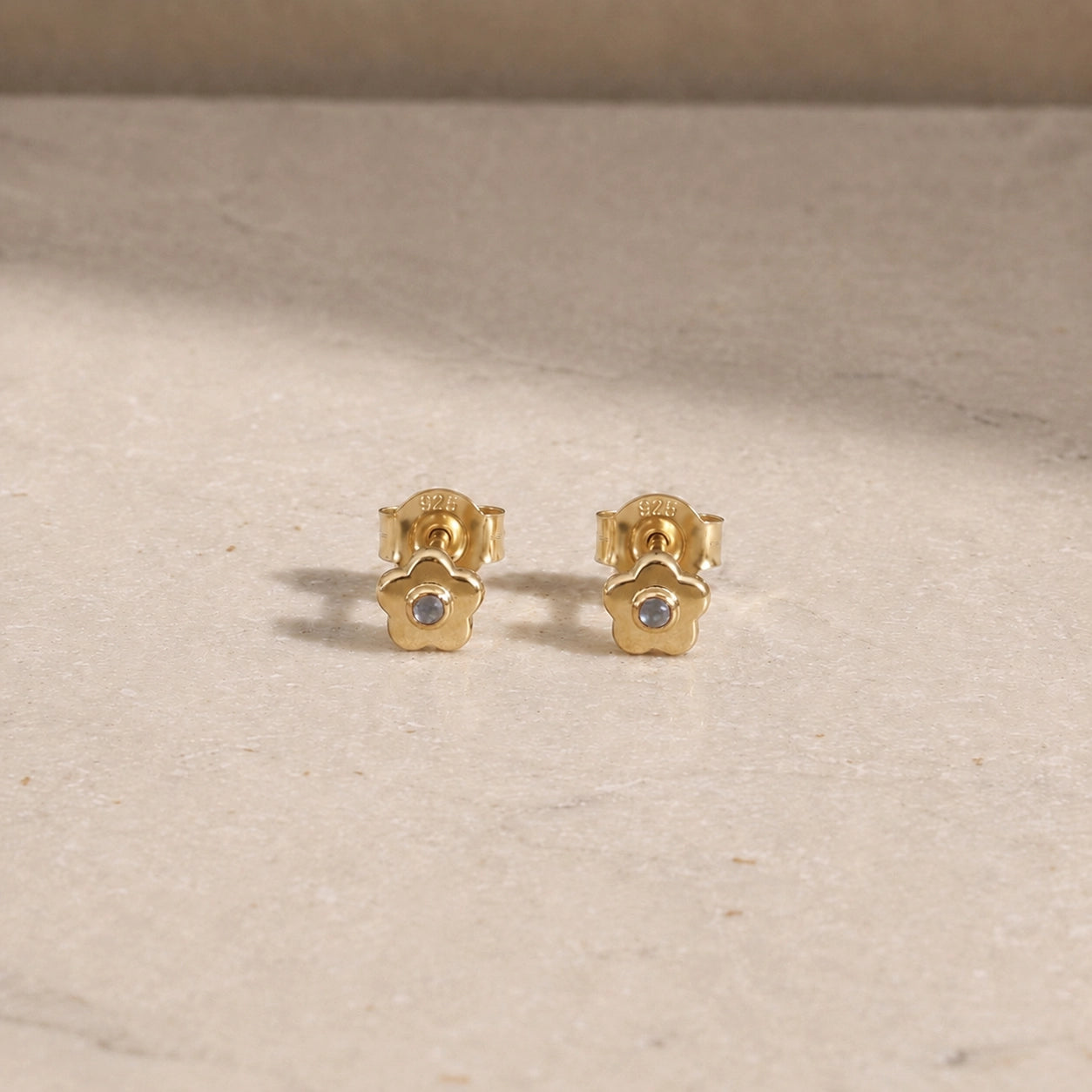 Pair of 14ct gold vermeil flower stud earrings with rainbow moonstone centre stones, displayed on a light beige stone surface with 925 butterfly backs visible.