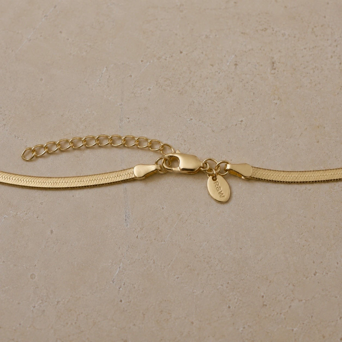 Close-up of a 14ct gold vermeil snake chain necklace featuring a lobster clasp, extender chain and Rose & Wolf branded tag on a neutral stone background.