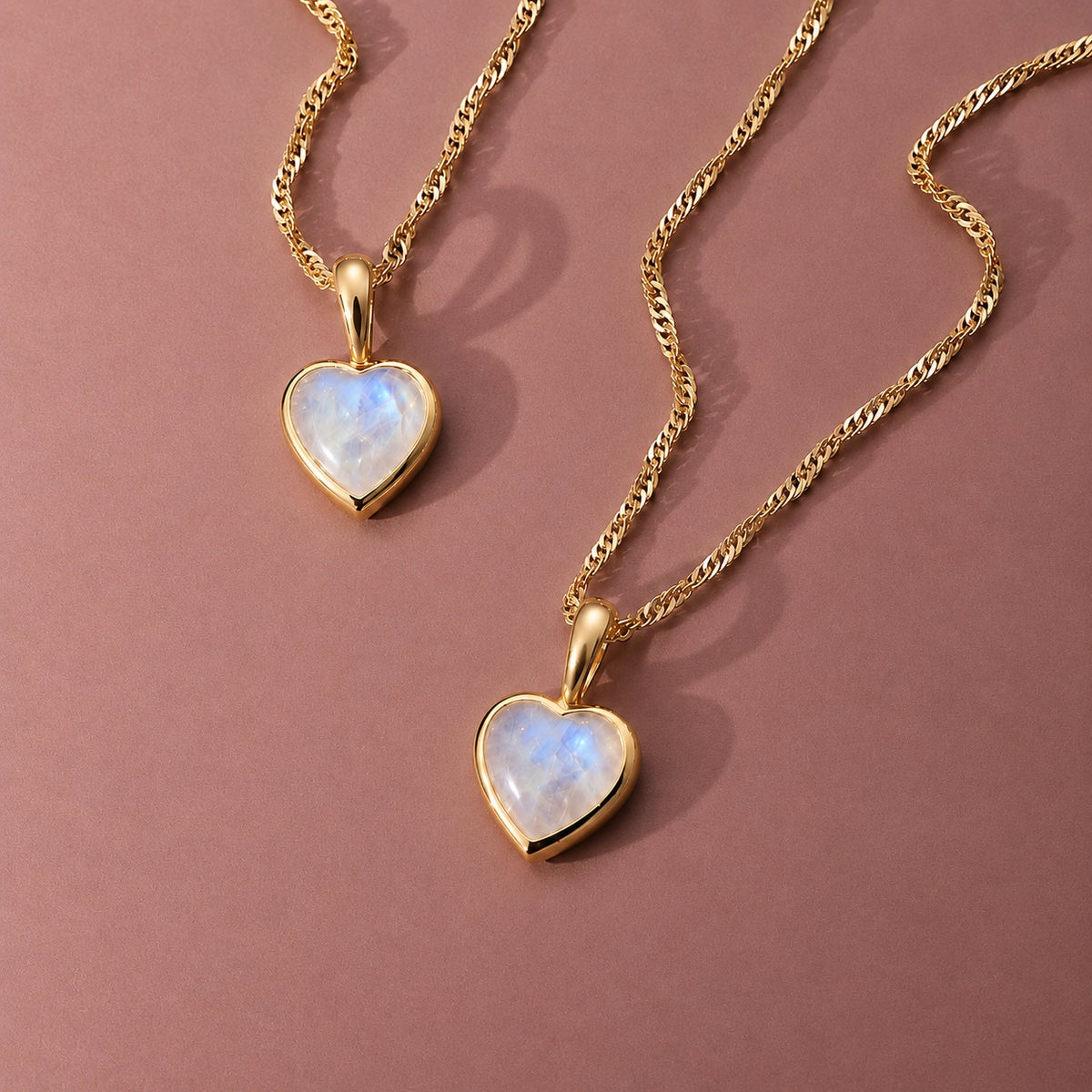 Two gold vermeil rainbow moonstone heart necklaces on fine Singapore chains, styled on a dusky pink background.