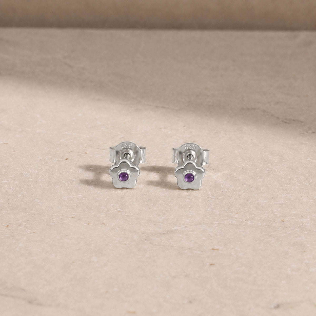 Pair of sterling silver flower stud earrings with amethyst centre stones, displayed on a light beige stone surface with 925 butterfly backs visible.