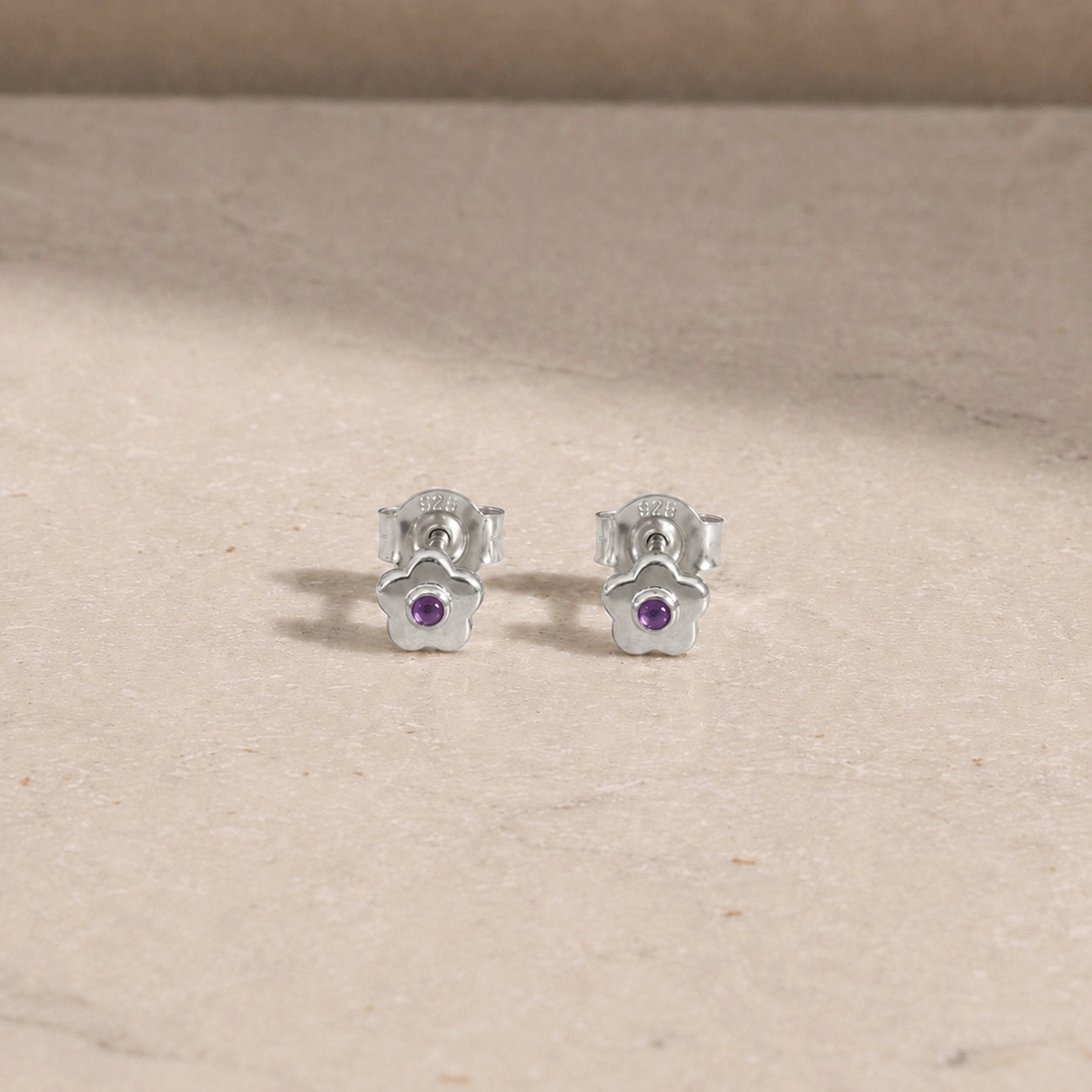 Pair of sterling silver flower stud earrings with amethyst centre stones, displayed on a light beige stone surface with 925 butterfly backs visible.