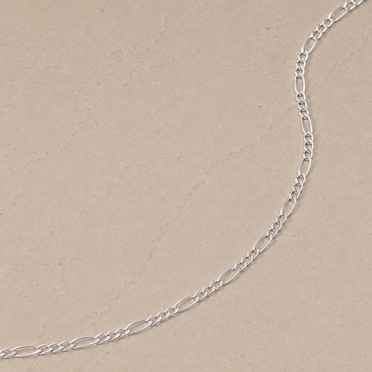 Close-up of a sterling silver Figaro chain necklace with alternating long and short links on a neutral beige stone background.