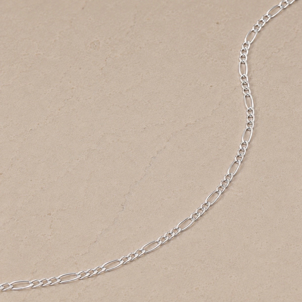 Close-up of a sterling silver Figaro chain necklace with alternating long and short links on a neutral beige stone background.