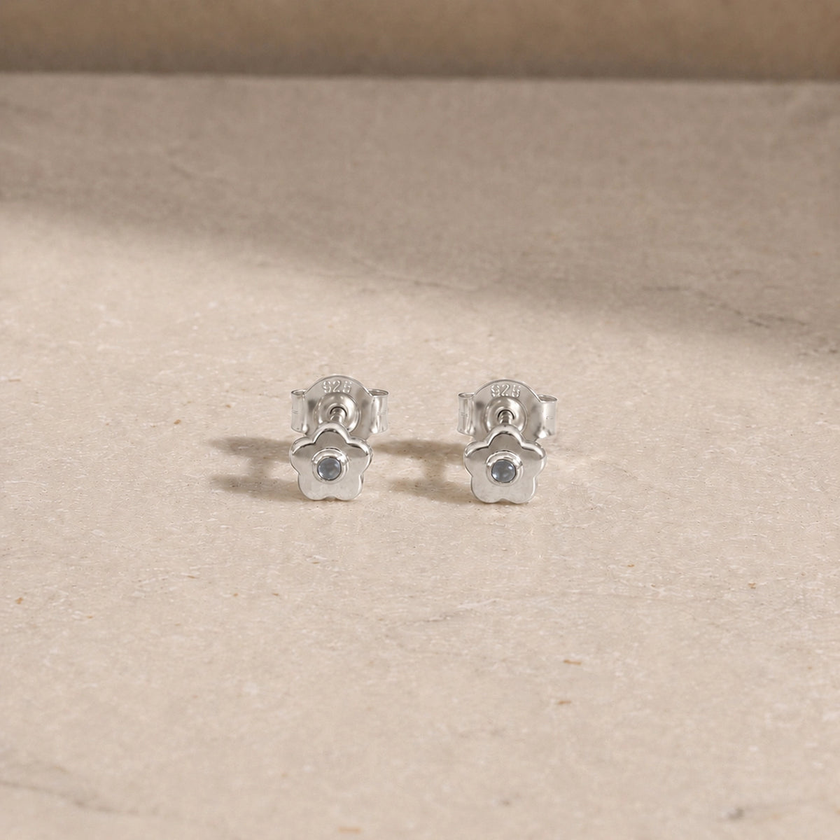 Pair of sterling silver flower stud earrings with rainbow moonstone centre stones, displayed on a light beige stone surface with 925 butterfly backs visible.
