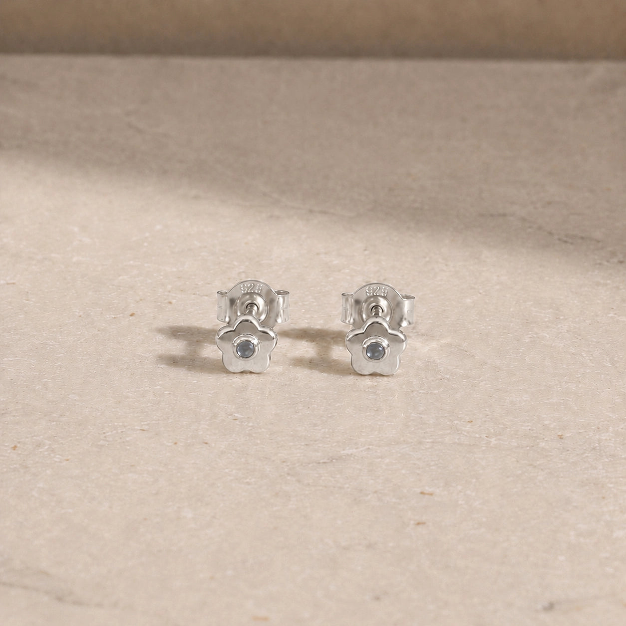 Pair of sterling silver flower stud earrings with rainbow moonstone centre stones, displayed on a light beige stone surface with 925 butterfly backs visible.