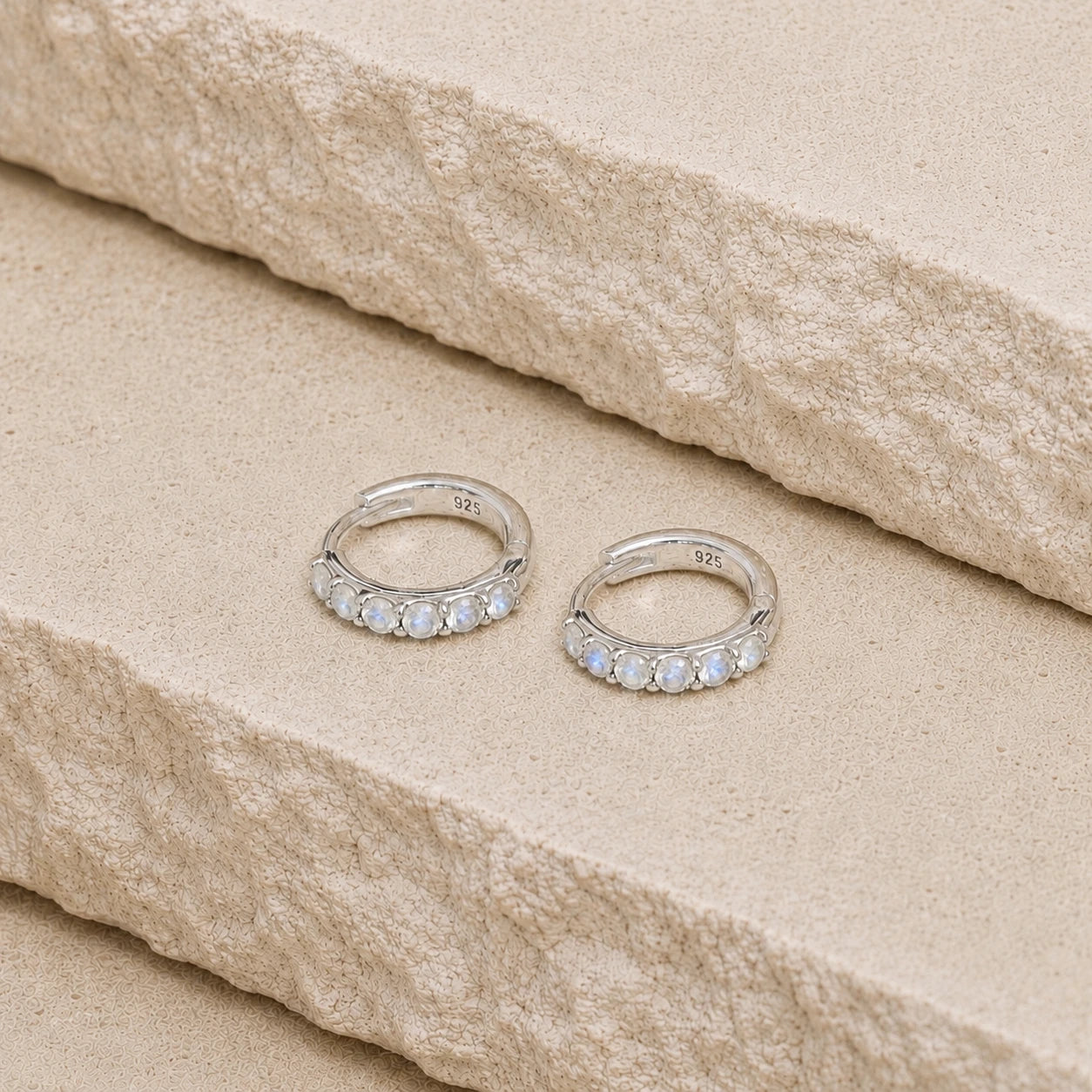 Sterling silver rainbow moonstone huggie hoop earrings with five light blue gemstones on each hoop, displayed on a textured cream stone surface with visible 925 hallmark inside each earring.