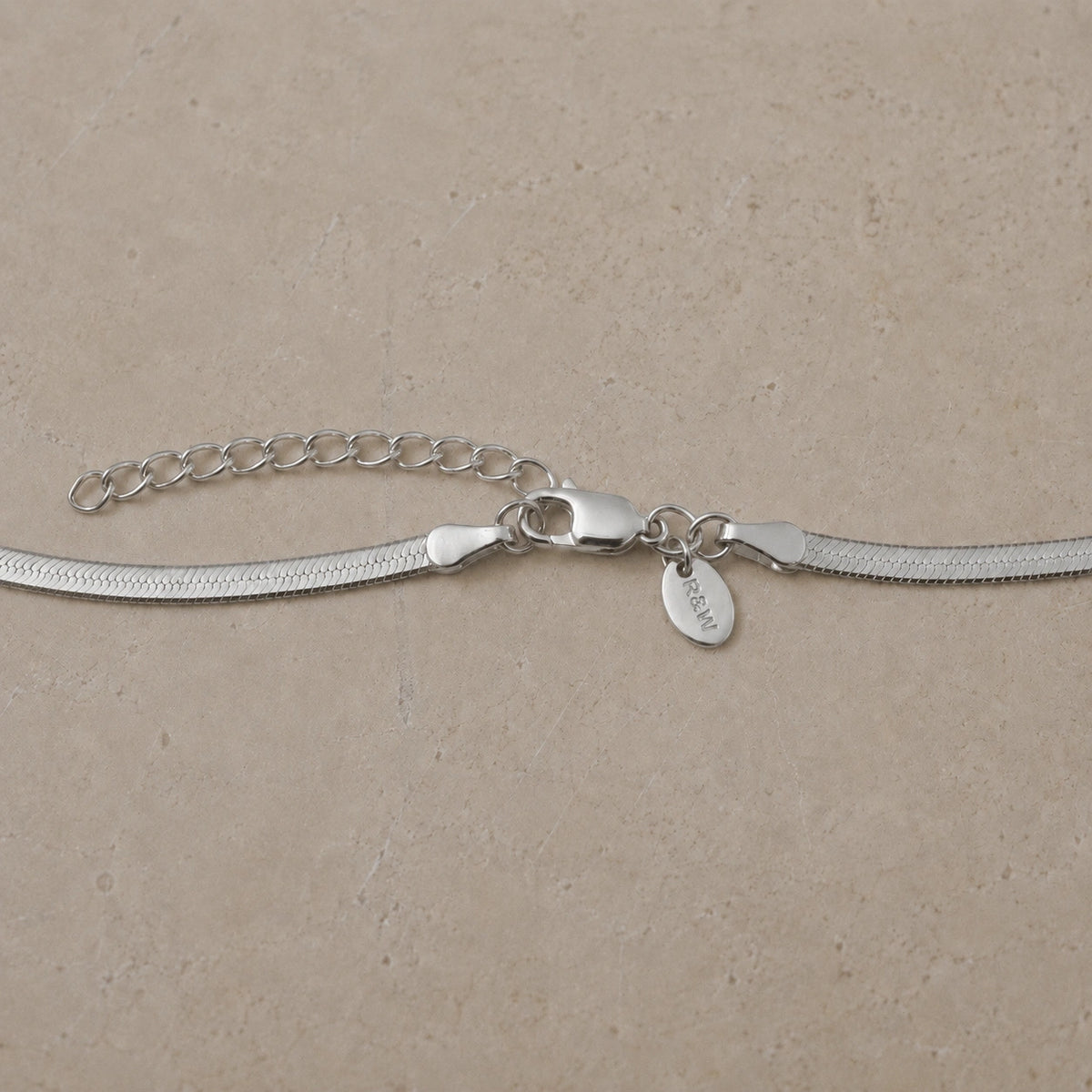 Close-up of a sterling silver snake chain necklace with lobster clasp, extender chain and branded charm tag on a neutral stone background.