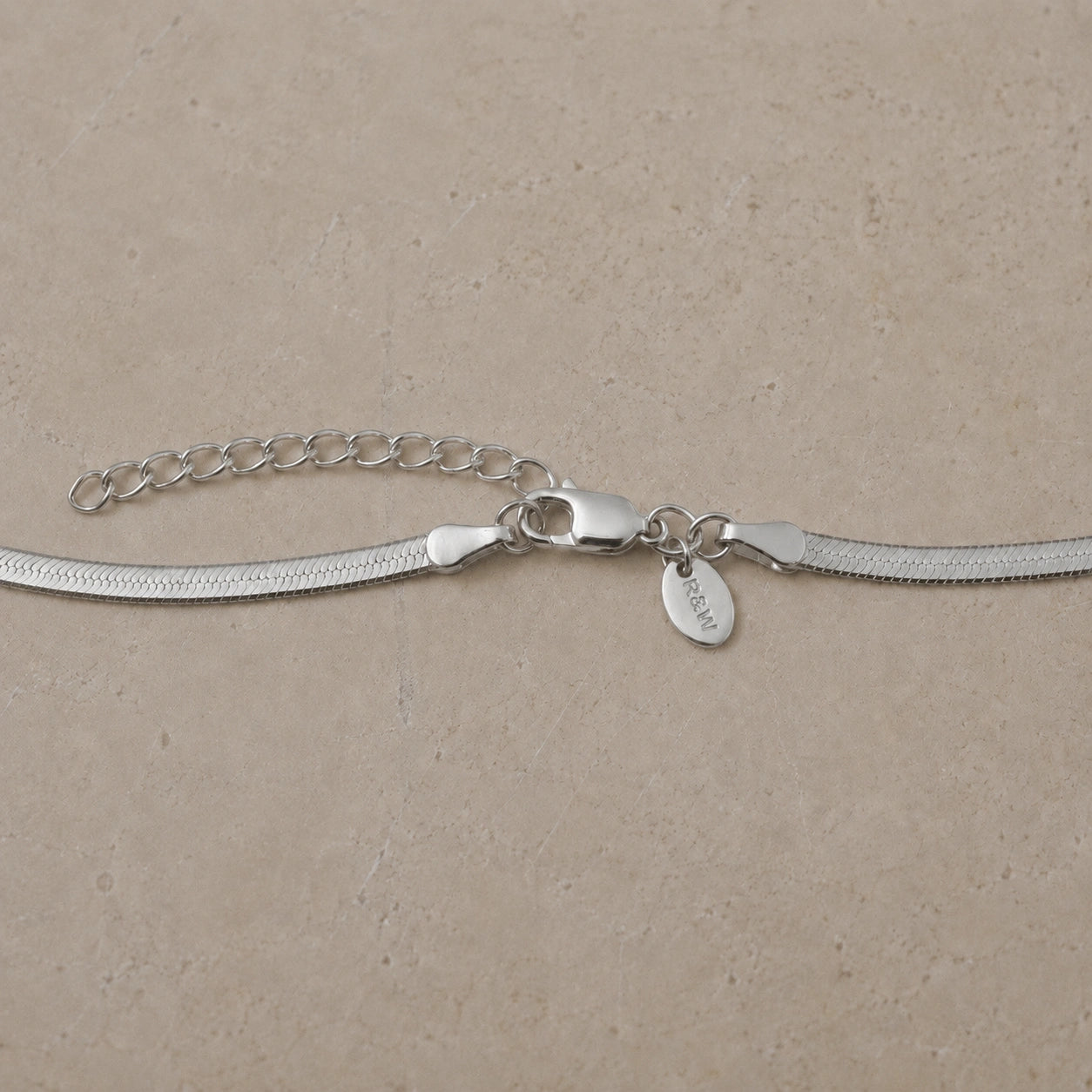Close-up of a sterling silver snake chain necklace with lobster clasp, extender chain and branded charm tag on a neutral stone background.